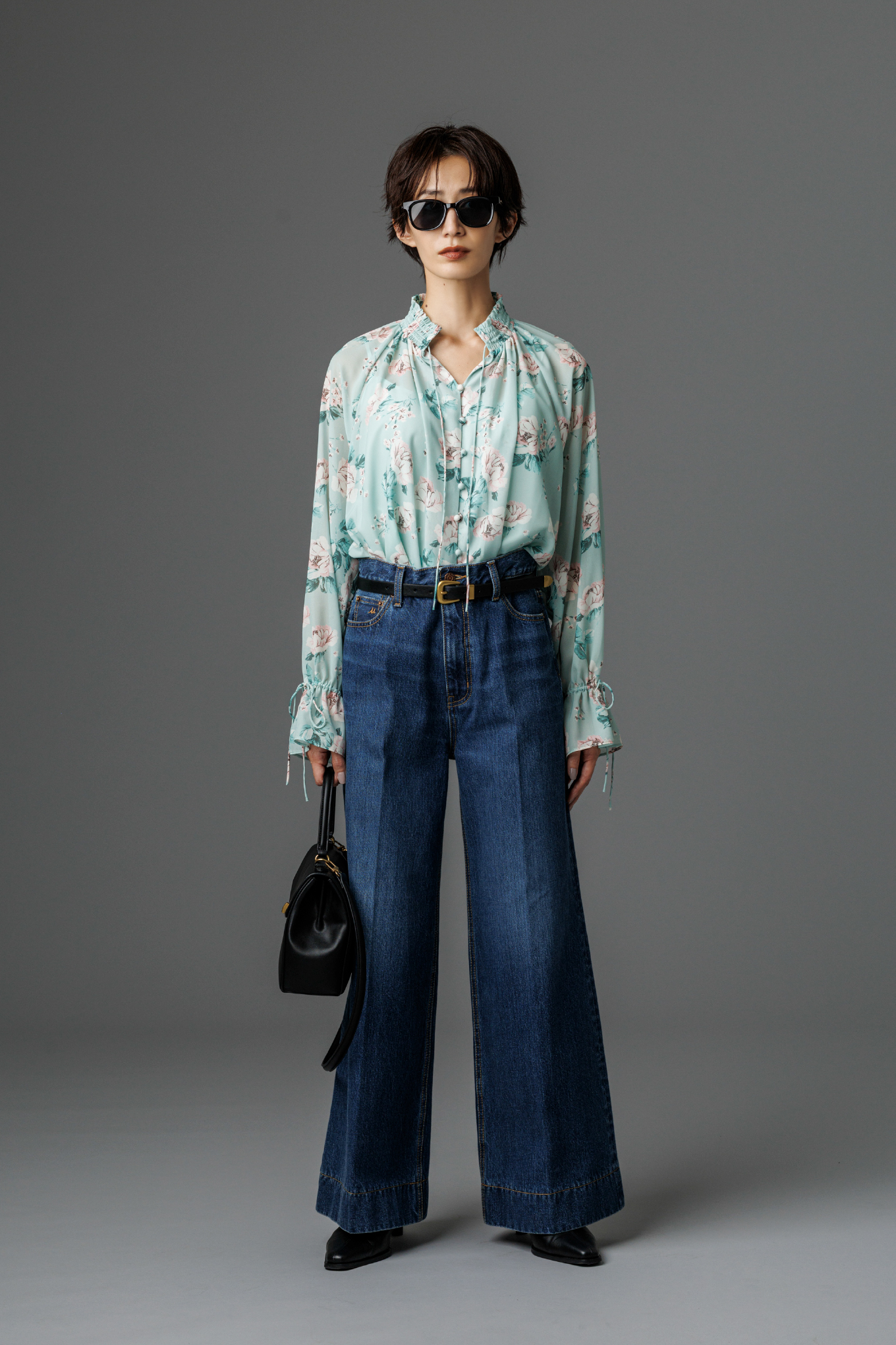 フラワープリントブラウス・ヘムデザインセミフレアデニムパンツ・メタルポイントセルフレームサングラス・ヴィンテージスリムベルト・メタルモチーフスクエアバッグ・キトゥンヒールショートブーツを着用した女性