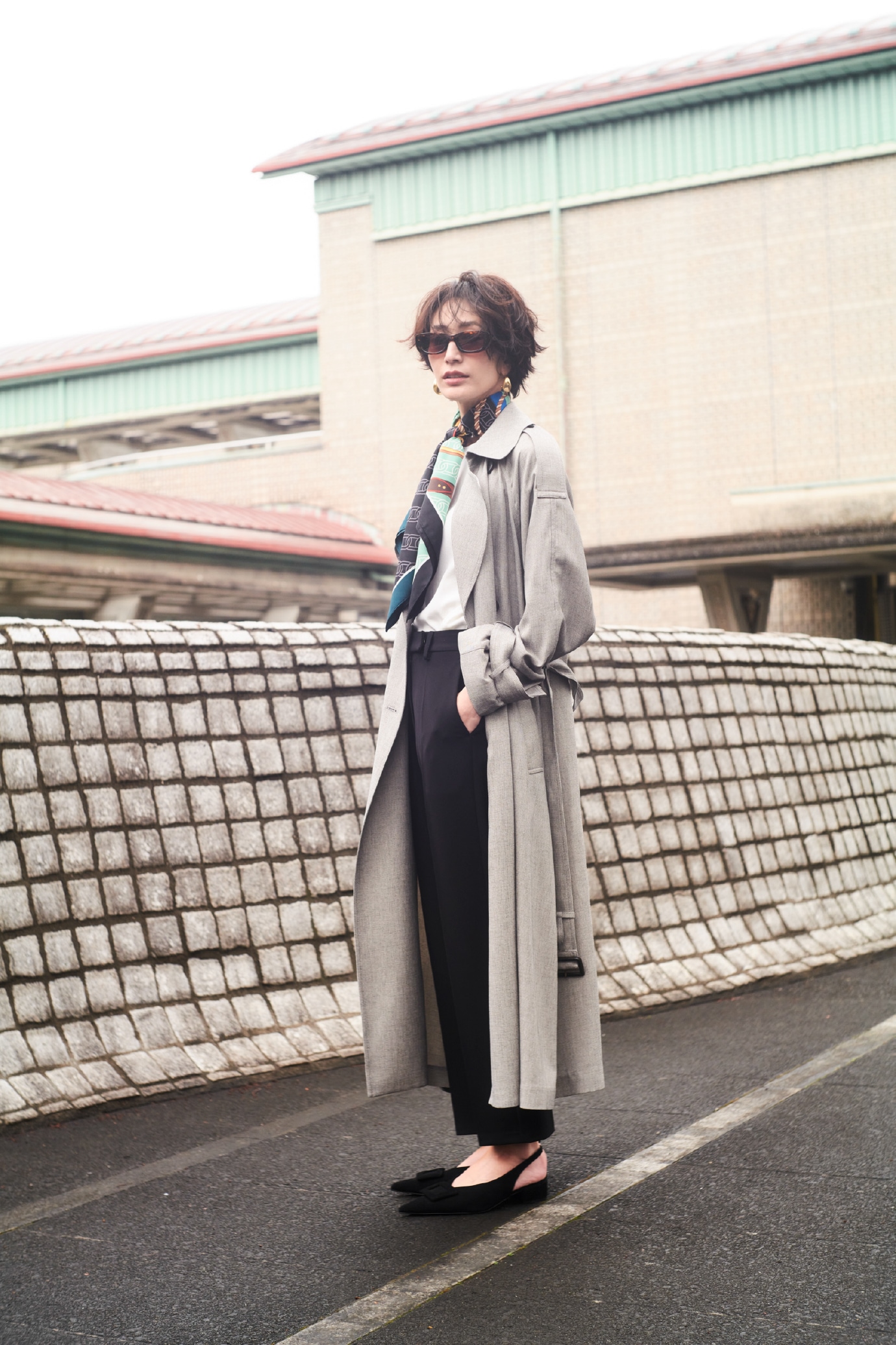 ミックスの撥水オーバーサイズトレンチコート・白のウォッシャブルニットトップス・黒の４サイズパンツとバックルモチーフバケットバッグ・ブラウンのセルフレームスクエアサングラス・ゴールドのメタルドロップピアス・グリーンのベルトモチーフ柄スカーフ・黒のバックル付きラムレザースリングパンプスを合わせた、媚びない大人の強さを感じさせるコーディネート