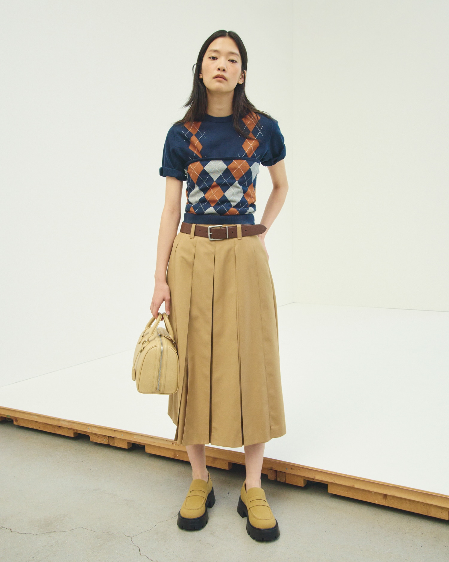 ネイビーのアーガイルニットトップスとアーガイルニットチューブトップ・キャメルのプリーツタックスカート・ダークブラウンのスクエアメタルサルカンベルト・ベージュのショルダー付きボストンバッグとモカシンローファーを合わせたコーディネート