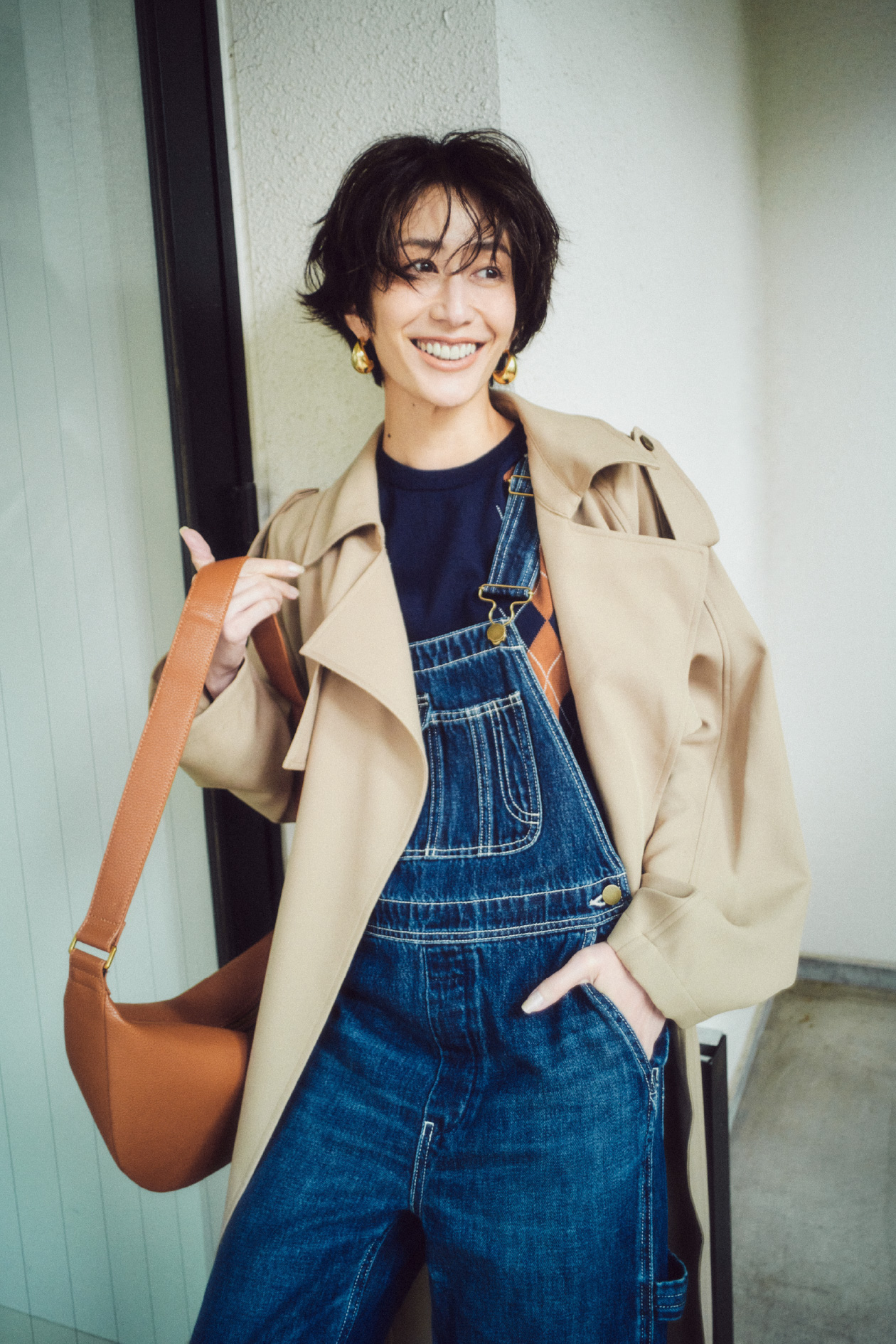 ベージュ色の撥水オーバーサイズトレンチコート、アーガイルニットトップス、デニムオーバーオール、メタルドロップピアス、ボディバッグを着用したモデル画像