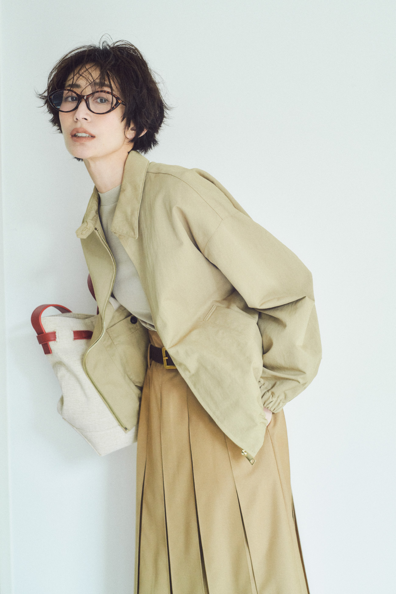 ベージュ色のフォルムデザインスウィングトップ、ウールテンセルハーフスリーブシアーＴ、プリーツタックスカート、キャットアイダテメガネ、スクエアバックルベルト、ベルト付きキャンバストートバッグを着用したモデル画像